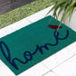 Lifestyle view of teal coir doormat with cursive navy 'home' and red bird on white tile by palm