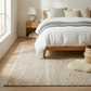 Lifestyle view of sand woven indoor outdoor rug beside bed with sheepskin throw