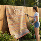 Woman hosing down multicolor striped outdoor rug hung on a tall wooden fence