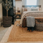 Lifestyle view of natural braided jute area rug with ivory border under bed and bench