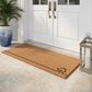 Lifestyle view of long natural coir doormat with black line-drawn house outline by lantern and palm