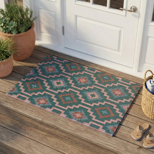 Lifestyle view of green and pink geometric woven doormat at white front door on a wooden porch