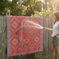 Woman rinsing orange and violet diamond outdoor rug hung on a wooden fence with a hose